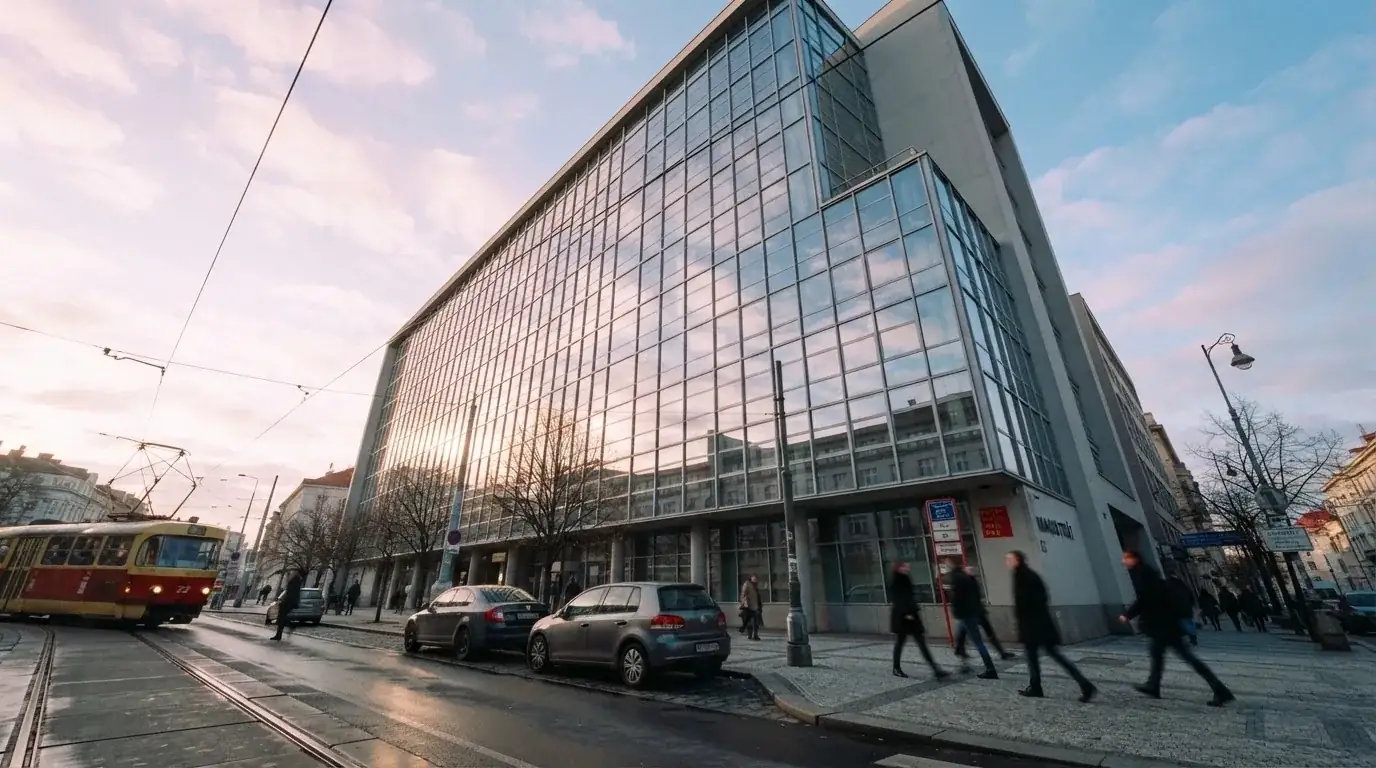 Fasáda moderní administrativní budovy s skleněnými okny odráží ranní oblohu nad rušnou pražskou ulicí.